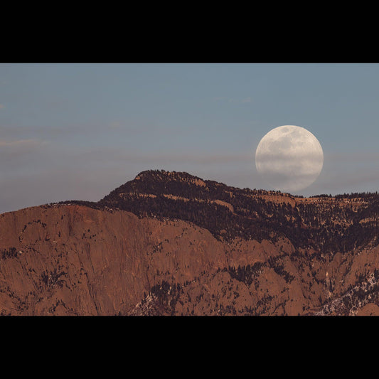 A full moon rising over the Sandia Mountains with a few clouds in front of it.